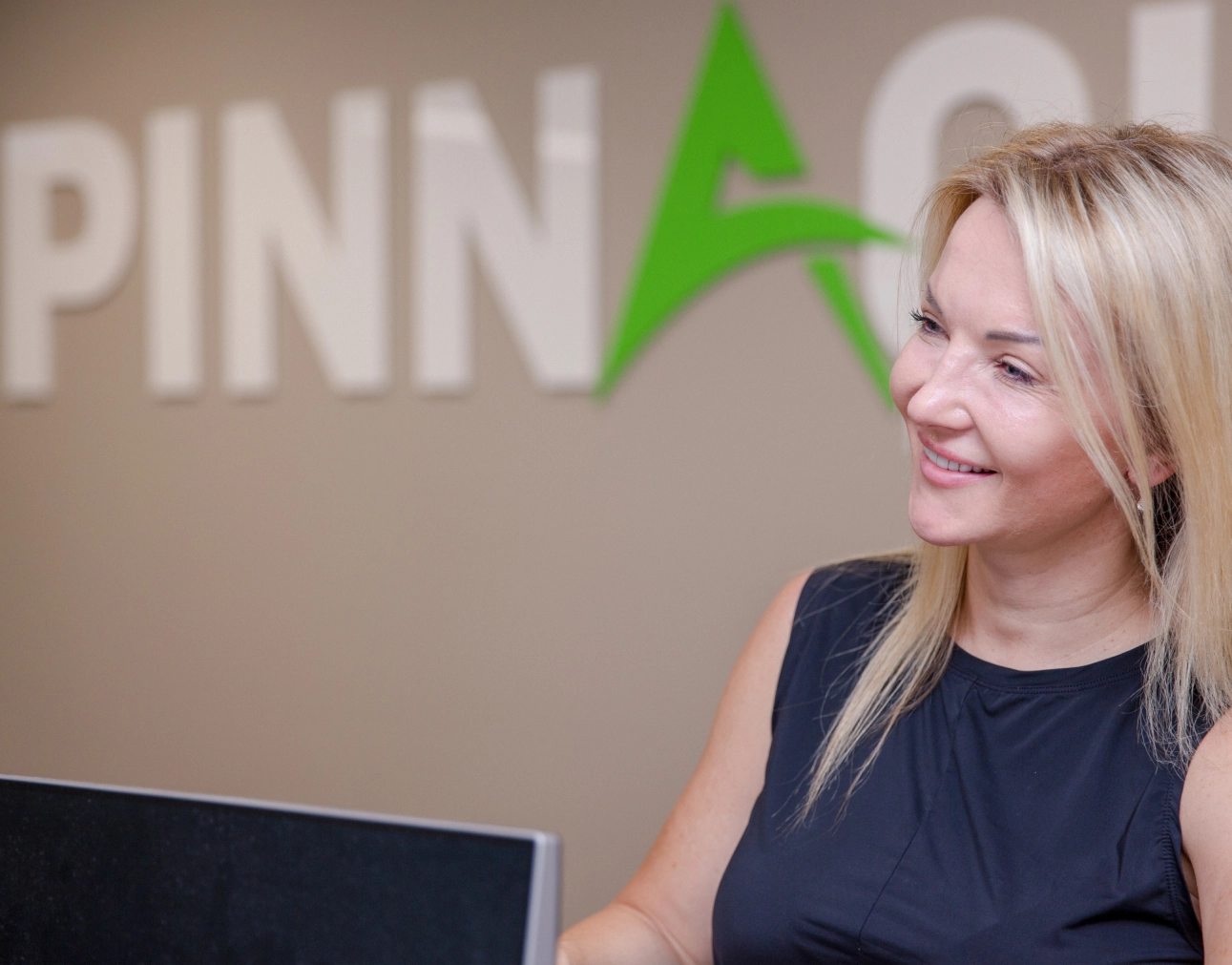 A woman with blonde hair smiles while standing near a computer monitor. The background features the partial word "PINN" and a green triangular logo, hinting at the Pinnacle brand.