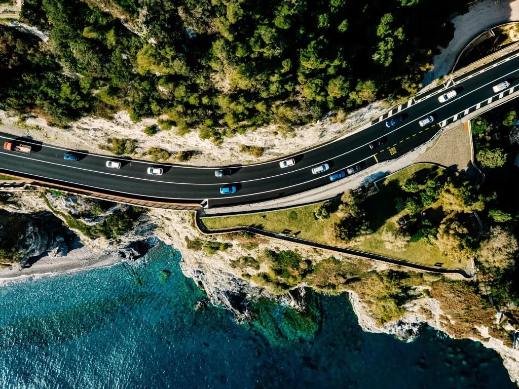 Aerial view of a winding coastal road with cars, bordered by turquoise waters on one side and dense green trees on the other.